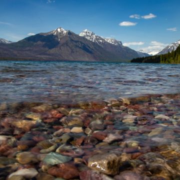 View of pebbles under the clear water of a mountain lake with snow-capped peaks and trees in the background