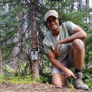 Headshot of Research Fellow Sujay Singh next to a tree with a trail camera installed on its trunk.
