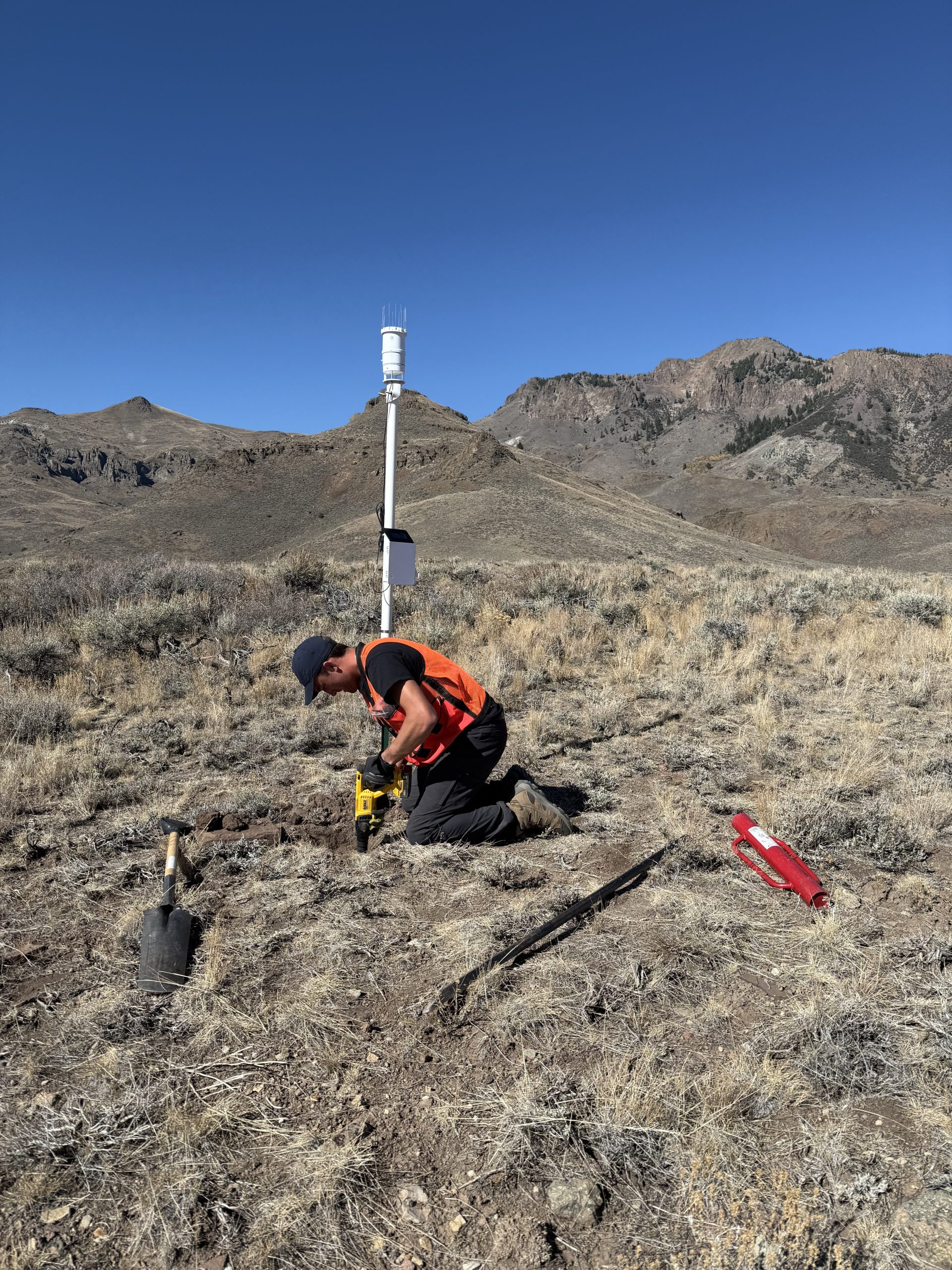 Researcher installs equipment for monitoring local conditions in a field of sagebrush with mountains in the distance.